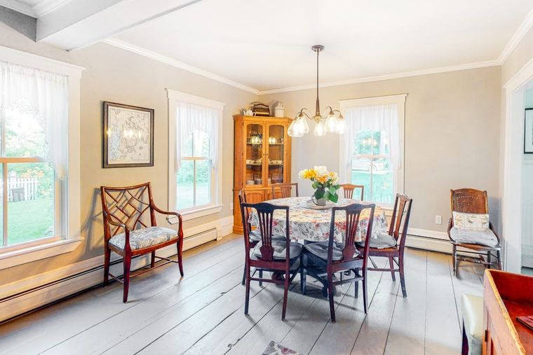 Sunlit dining room with a round table, antique china cabinet, and painted plank floors.