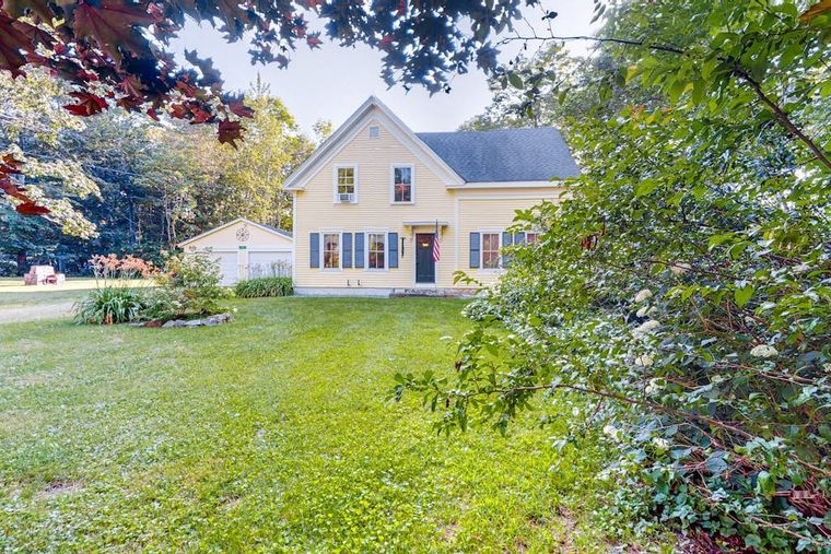 Front of the pale-yellow farmhouse with blue shutters and autumn trees in Penobscot, Maine.