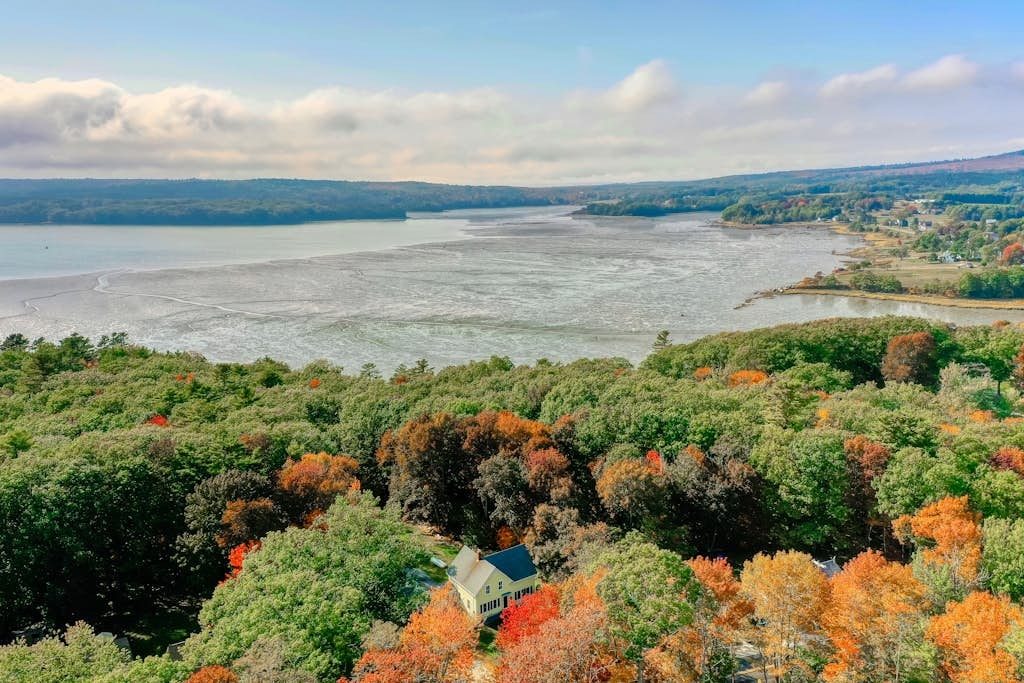 Aerial view of the buttercream Farmhouse on the Cove tucked among autumn trees above a wide tidal cove of Penobscot Bay in Penobscot, Maine.